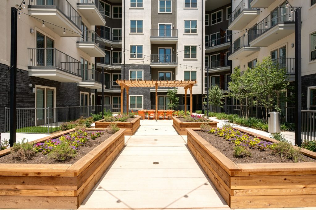 view from courtyard planters of outdoor area with large wooden table, seating, and wood pergola